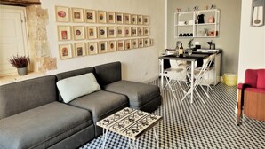 Ein Wohnzimmer mit grauem Sofa, einem Couchtisch mit Muster, einer Wand voller gerahmter Bilder, einer kleinen Küchenzeile mit weißem Regal und einem gemusterten Boden.
