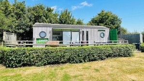 Une petite construction en bois d’un seul étage, avec une porte et des fenêtres vertes, entourée d’une terrasse en bois et d’une haie.