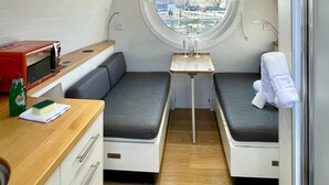 A compact kitchen with a microwave, a sink, and a small table. There are two seating areas with cushions and a window providing a view outside.