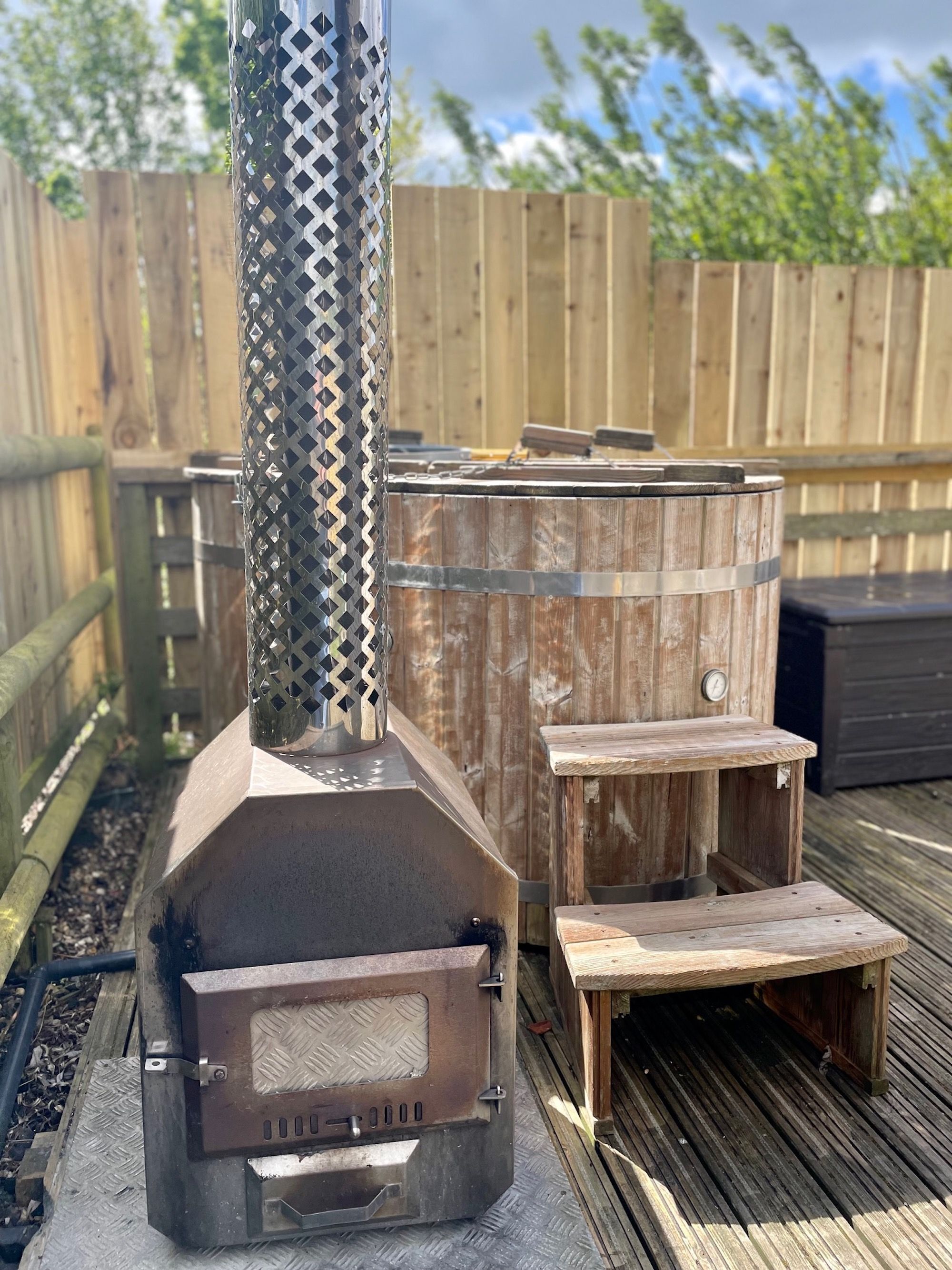 Un poêle à bois portatif, doté d’une grille en métal, accompagné d’un banc en bois et d’une petite table en bois, le tout installé sur une terrasse en bois.