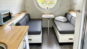 A compact, well-lit room with a kitchenette, a small table with a lamp, and seating areas with cushions.