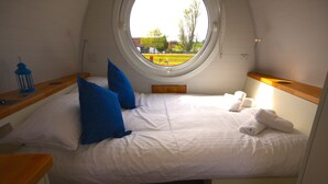 A room with a bed, two blue pillows, a wooden bedside table with a blue lantern, and a round window with a view of greenery outside.