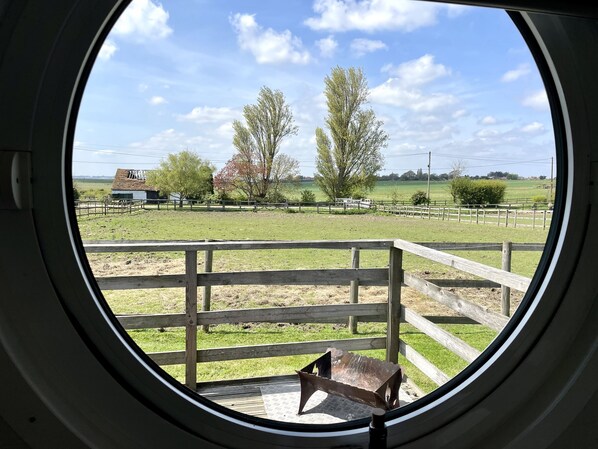 Une vue à travers une fenêtre circulaire sur un champ verdoyant, avec une clôture en bois, un petit bâtiment et des arbres.