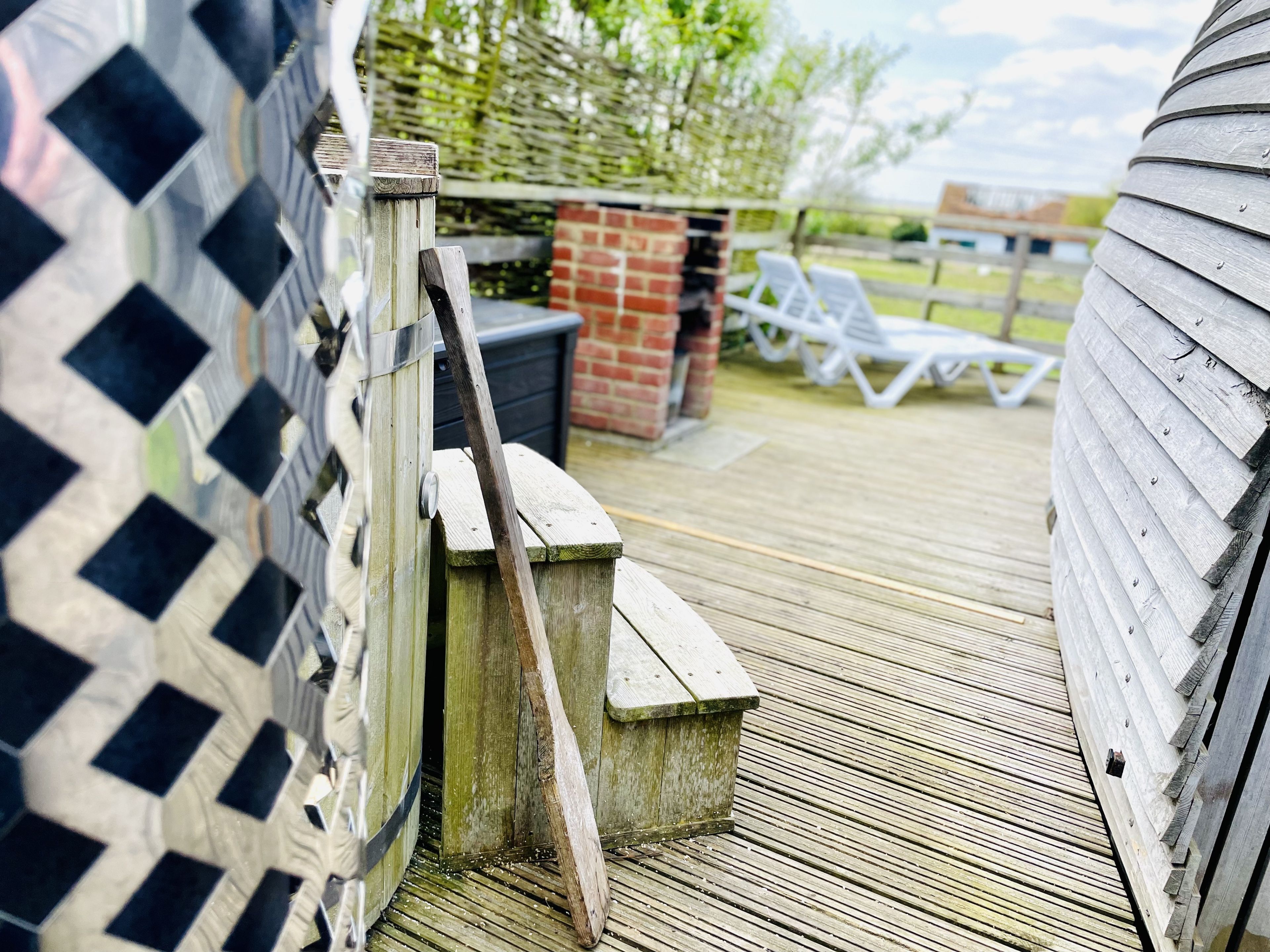 Une terrasse en bois avec une porte en métal, une structure en briques et des chaises longues blanches.