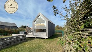 Una pequeña estructura en forma de A de madera, con un amplio ventanal, rodeada por una cerca de madera y follaje.