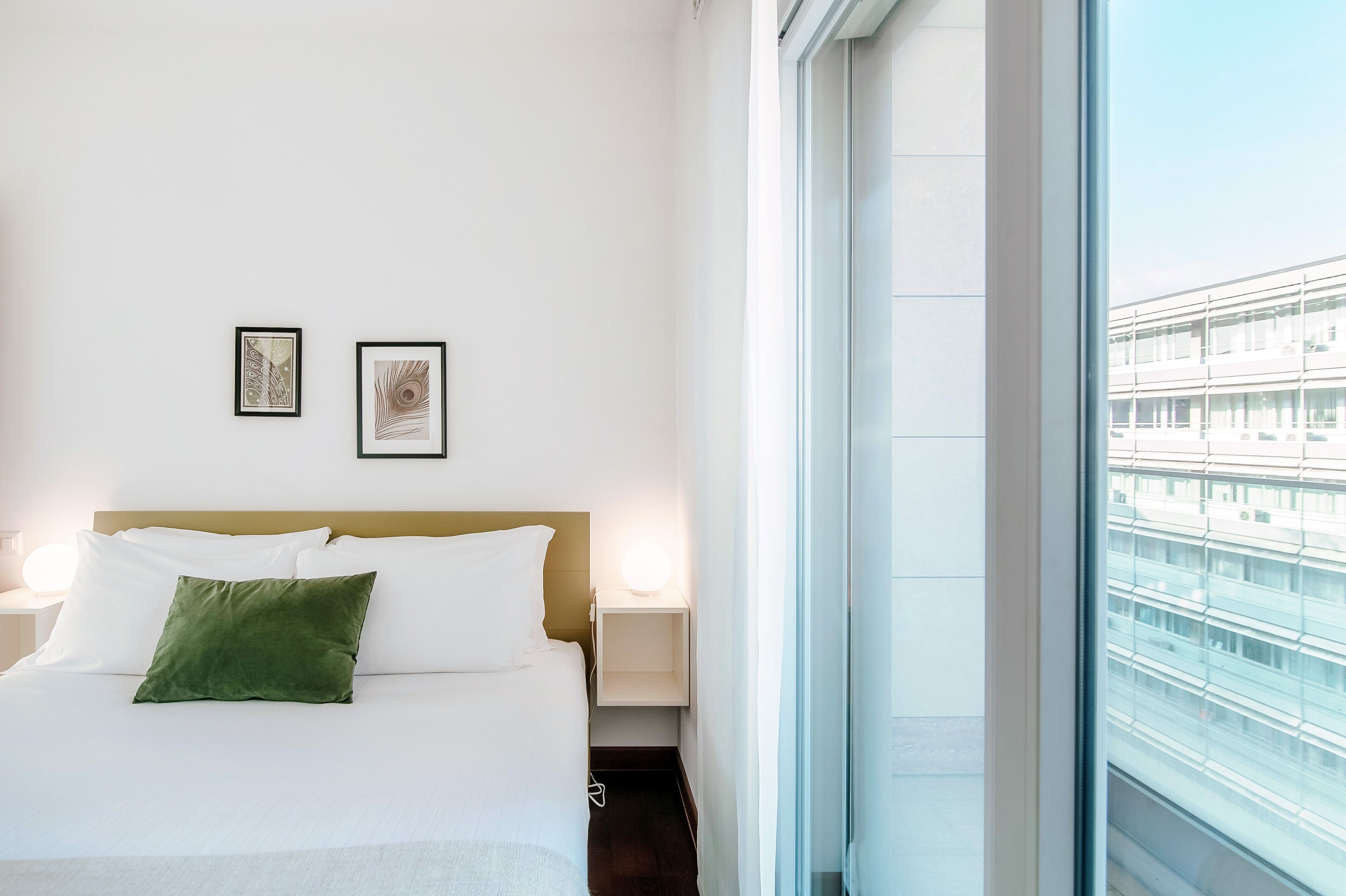 Una habitación de hotel con una cama, dos cuadros en la pared, una lámpara de noche y una ventana con vistas a un edificio.