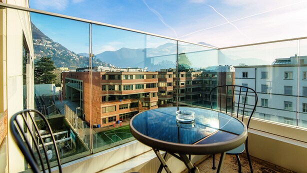 Un balcon doté d’une table et de chaises en verre, offrant une vue sur un paysage urbain avec des bâtiments et des montagnes.