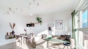 Un salon moderne avec un canapé, une table à manger et des chaises. On y trouve des plantes, des œuvres d’art et une fenêtre avec des rideaux.