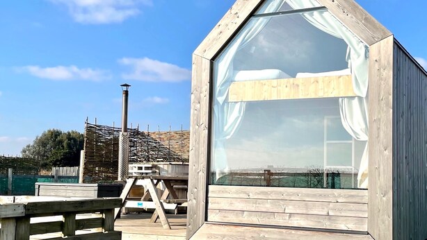 Eine Holzhütte mit Satteldach und großen Fenstern, auf einer Wiese mit Holzterrasse und Picknicktisch gelegen.