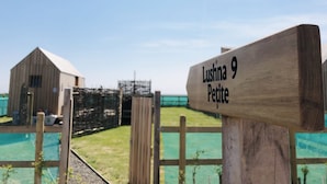 Ein hölzernes Schild mit der Aufschrift "Lushna 9 Petite", vor einem blauen Himmel und einem eingezäunten Bereich mit einem Gebäude in der Ferne.