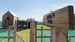 A wooden sign with "Lushna 9 Petite" written on it, set against a backdrop of a clear sky and a fenced area with a building in the distance. - Lushna Petite 8 at Lee Wick Farm Glamping (Clacton on Sea)