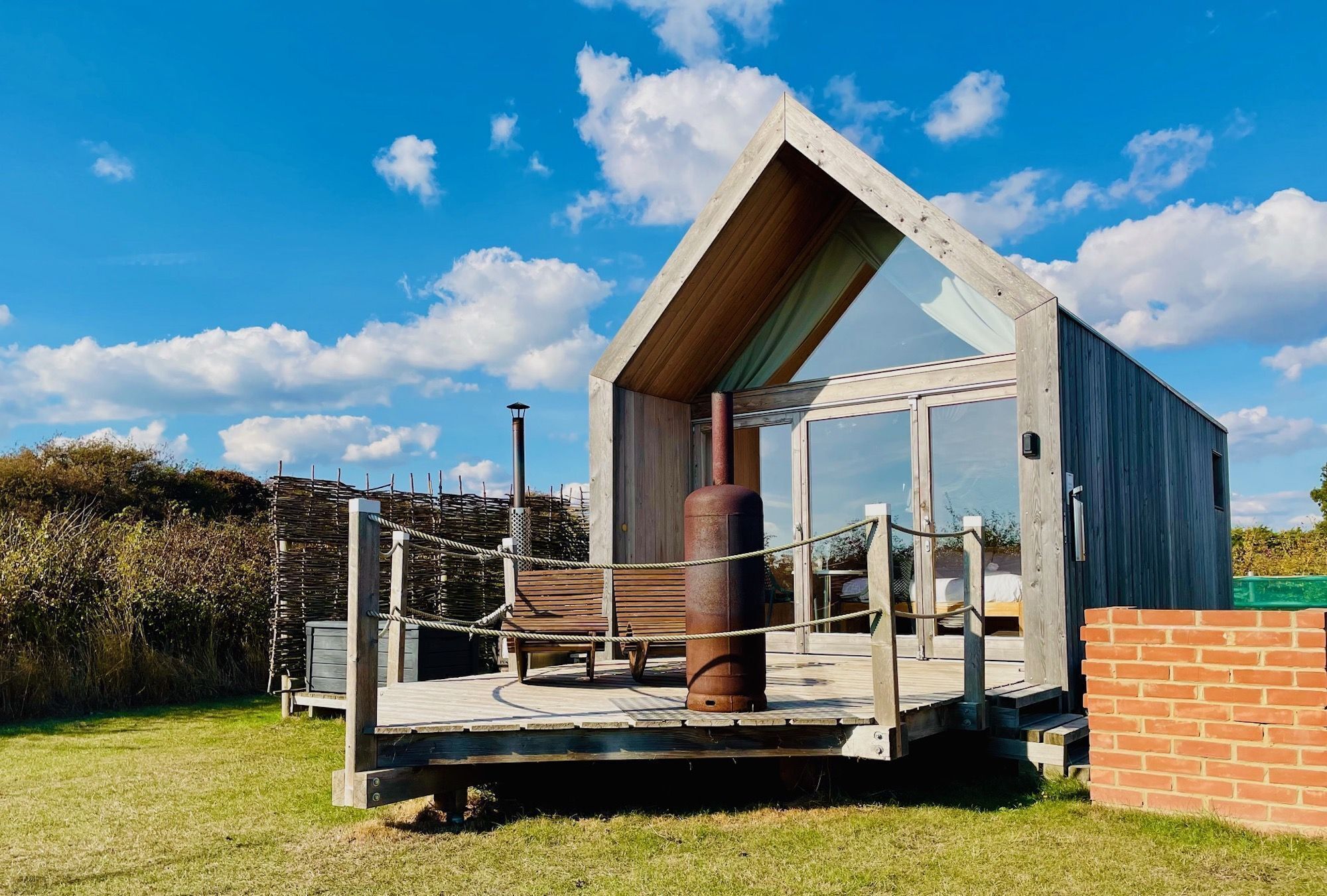 Une cabane moderne en bois, dotée d’une porte en verre, d’une terrasse en bois et d’une barrière en corde.