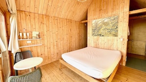 A wooden room with a bed, a small table, and a chair. There is a shelf with bottles and glasses, and a painting on the wall. - Lushna Petite 12 at Lee Wick Farm Glamping (Clacton on Sea)