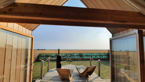 A wooden deck with two wooden chairs and a table, overlooking a beach with a fence and greenery. - Lushna Petite 12 at Lee Wick Farm Glamping (Clacton on Sea)