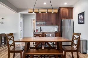 A modern kitchen with wooden cabinets, a stainless steel refrigerator, and a wooden dining table with chairs. - Luxury Boyne Mountain Ski-In/Out with Hot Tub (Boyne City)