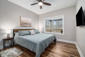 A bedroom with a bed, a ceiling fan, a window with a view of trees, a mounted TV, and a bedside table with a lamp. - Luxury Boyne Mountain Ski-In/Out with Hot Tub (Boyne City)
