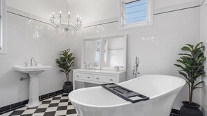 Une salle de bain avec une baignoire sur pieds, un lavabo sur colonne, une coiffeuse avec un miroir et un lustre.