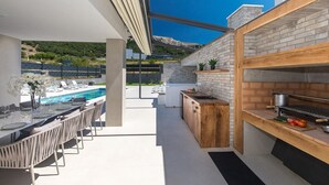 Une cuisine extérieure moderne avec vue sur la piscine, des meubles de rangement en bois et un coin repas avec une table dressée pour quatre personnes.