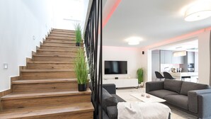 Un salon moderne avec un escalier en bois, des canapés gris, un téléviseur à écran plat et une cuisine en arrière-plan.