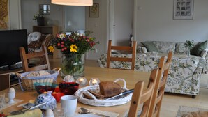 Ein Essbereich mit einem Tisch, der für ein Frühstück gedeckt ist, einschließlich Brot, Eier und Obst. Im Hintergrund befinden sich ein Fernseher, Stühle und ein floral gemustertes Sofa.