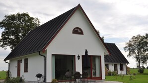 Ein weißes Gebäude mit schwarzem Dach und rotem Zierstreifen, ausgestattet mit einer großen Schiebeglastür und einer Terrasse mit Gartenmöbeln.