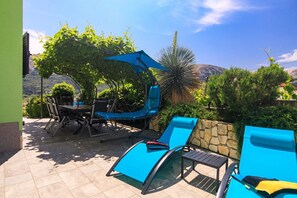 Pátio externo com mesa e cadeiras, um guarda-sol azul e área de piscina com espreguiçadeiras.