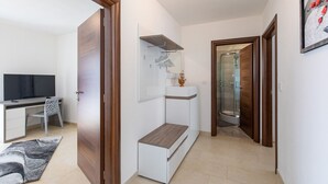A modern bedroom with a bed, bedside table, and a wardrobe. There is a desk with a chair and a TV in the adjacent room.