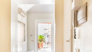 Un couloir doté d’un plancher en bois, une porte blanche avec une petite fenêtre, et une plante en pot visible à travers la fenêtre.