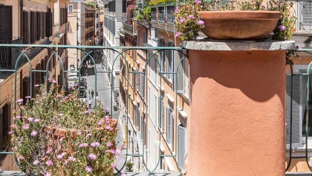 Une rue étroite bordée de bâtiments des deux côtés, un patio dallé avec deux tables de style lanterne, et un grand pot en terre cuite rempli de fleurs.