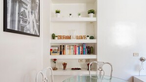 Un coin repas avec une table en verre, des chaises blanches, une bibliothèque remplie de livres et de plantes, et un tableau encadré au mur.