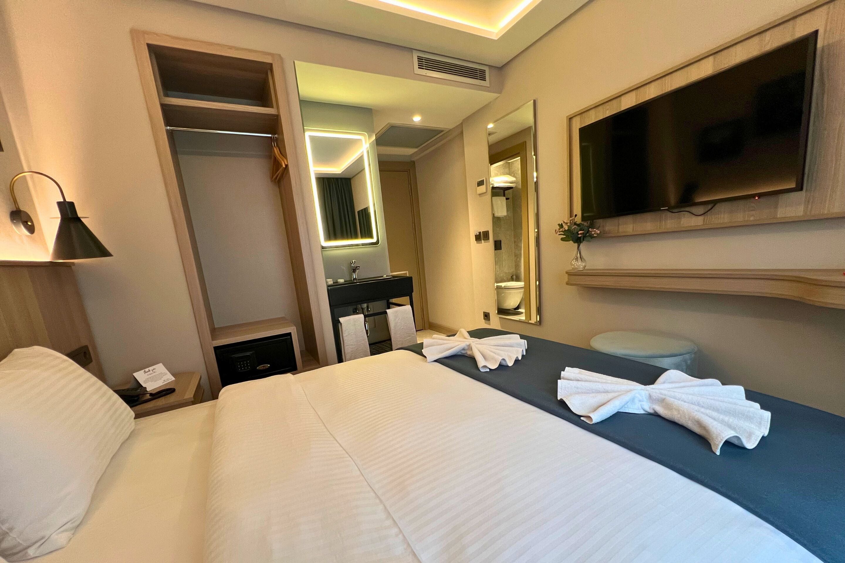 Une chambre d’hôtel comprenant un lit, une table de chevet avec une lampe, un miroir, un téléviseur fixé au mur, une salle de bain avec un lavabo et un miroir, et des serviettes sur le lit.