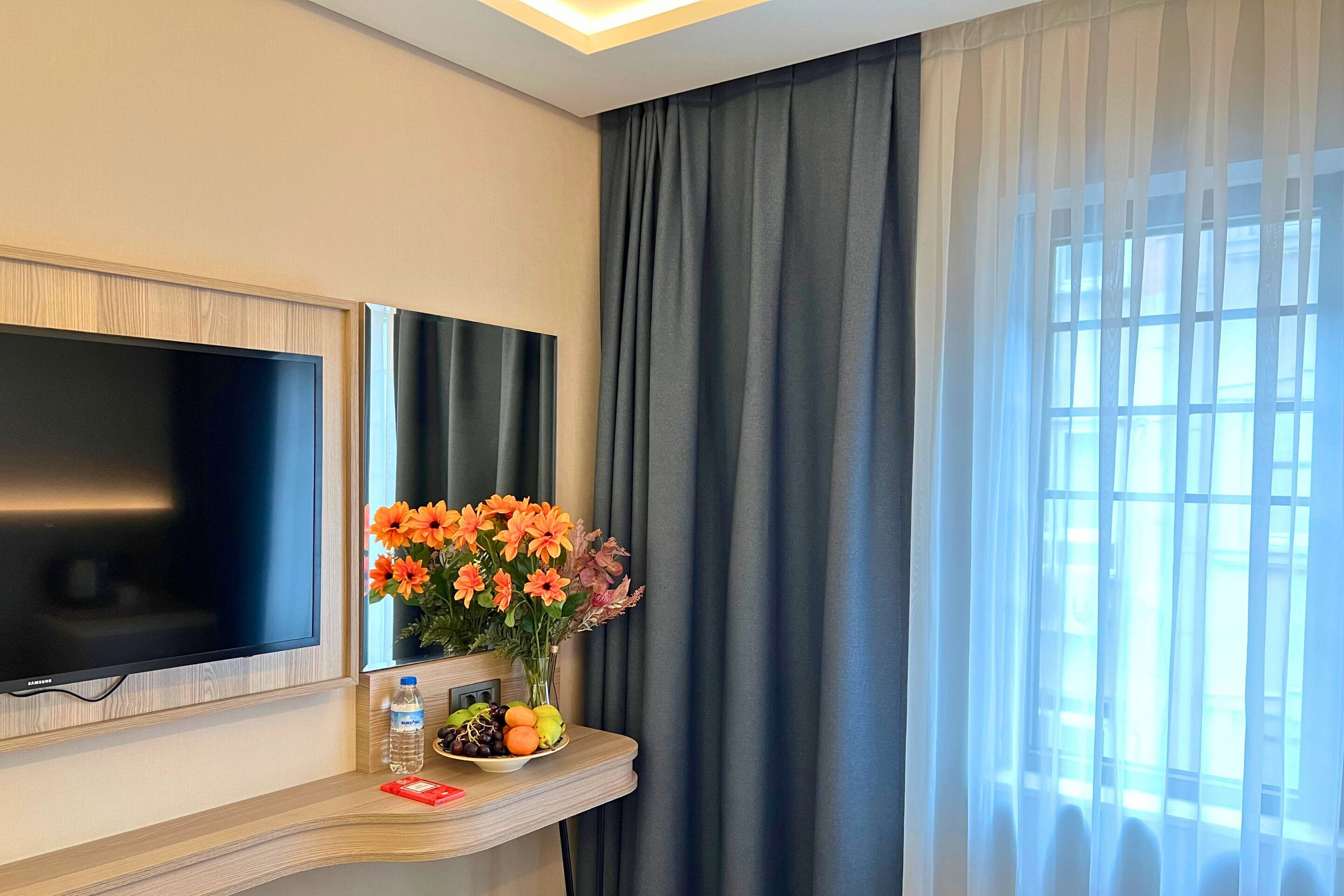 A hotel room with a flat-screen TV mounted on the wall, a small table with a vase of flowers, a fruit bowl, and a water bottle.