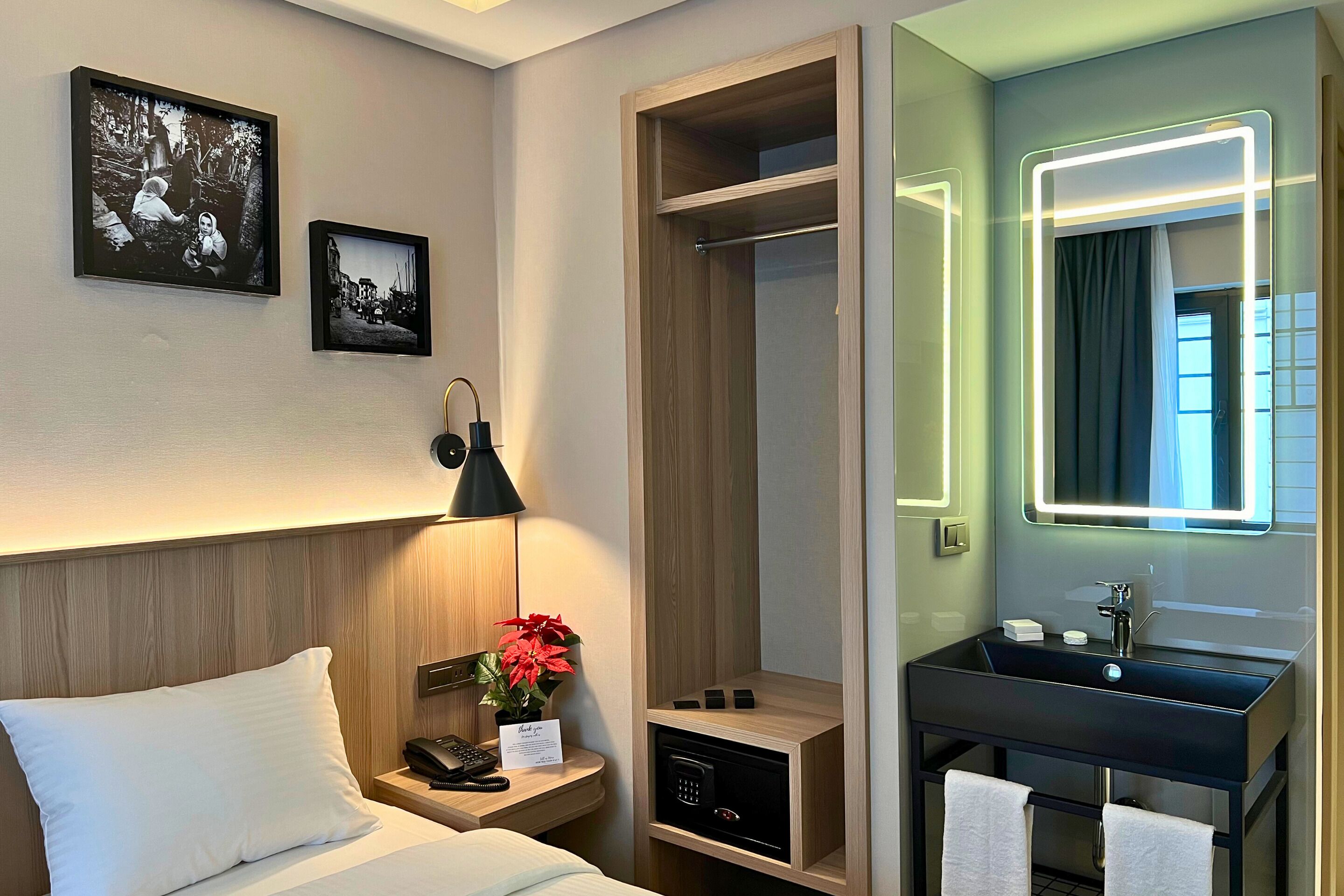 A hotel room with a bed, bedside table, lamp, framed pictures, a closet, and a bathroom with a sink and mirror.