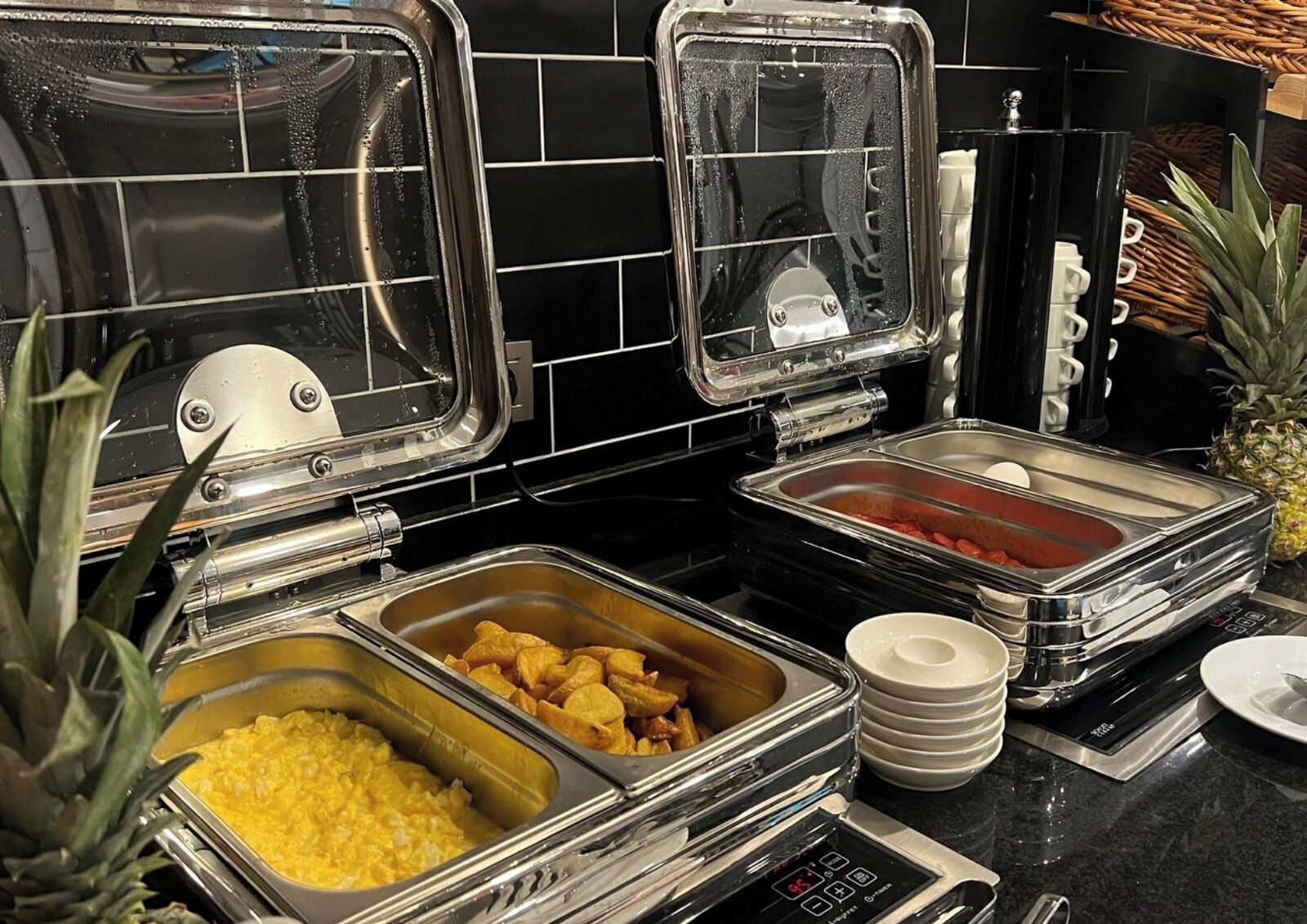 A buffet setup with various food items in metal containers, a coffee machine, and a stack of white plates.