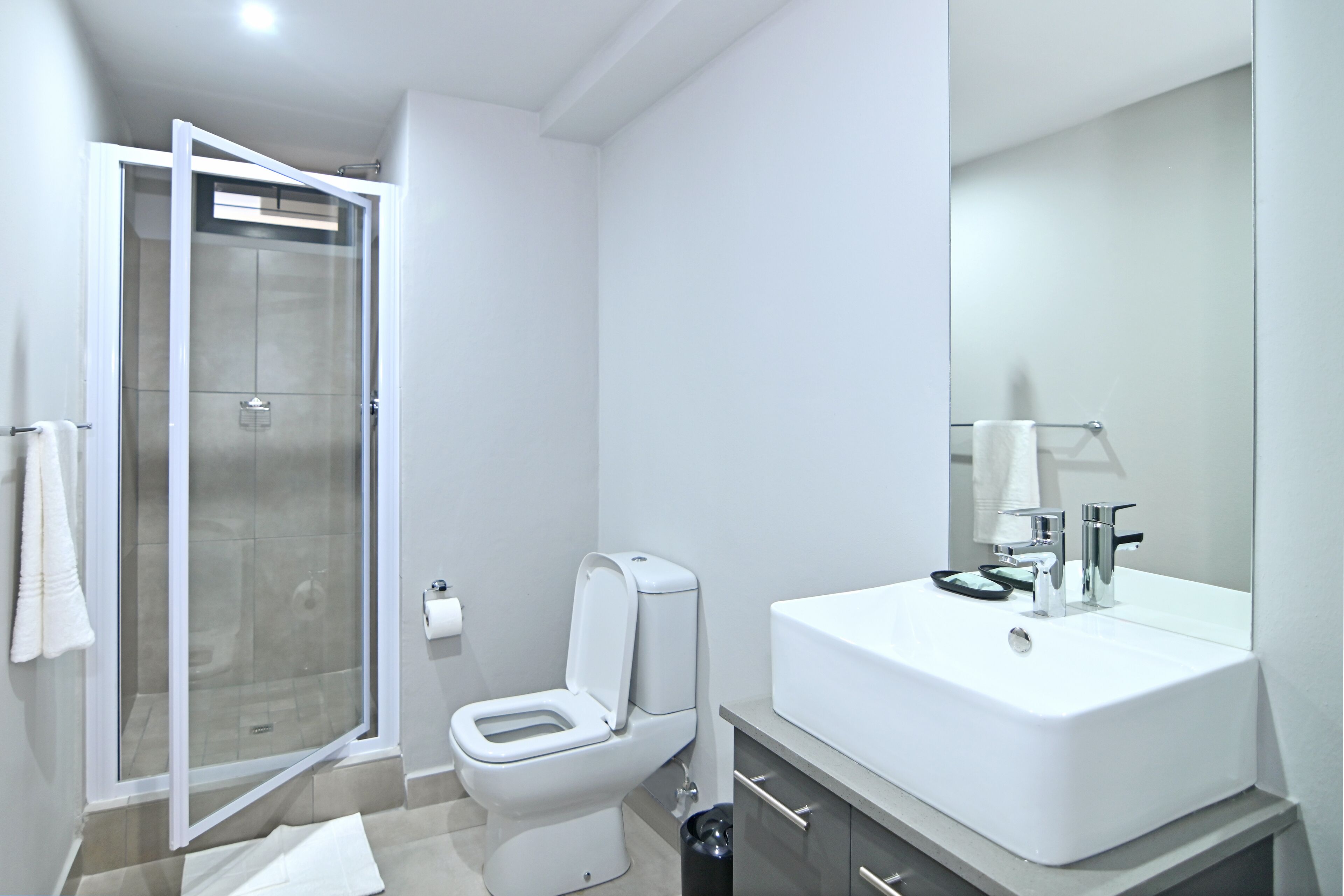 Un baño moderno con ducha de vidrio, inodoro blanco, un lavamanos grande con espejo y un pequeño cubo de basura.