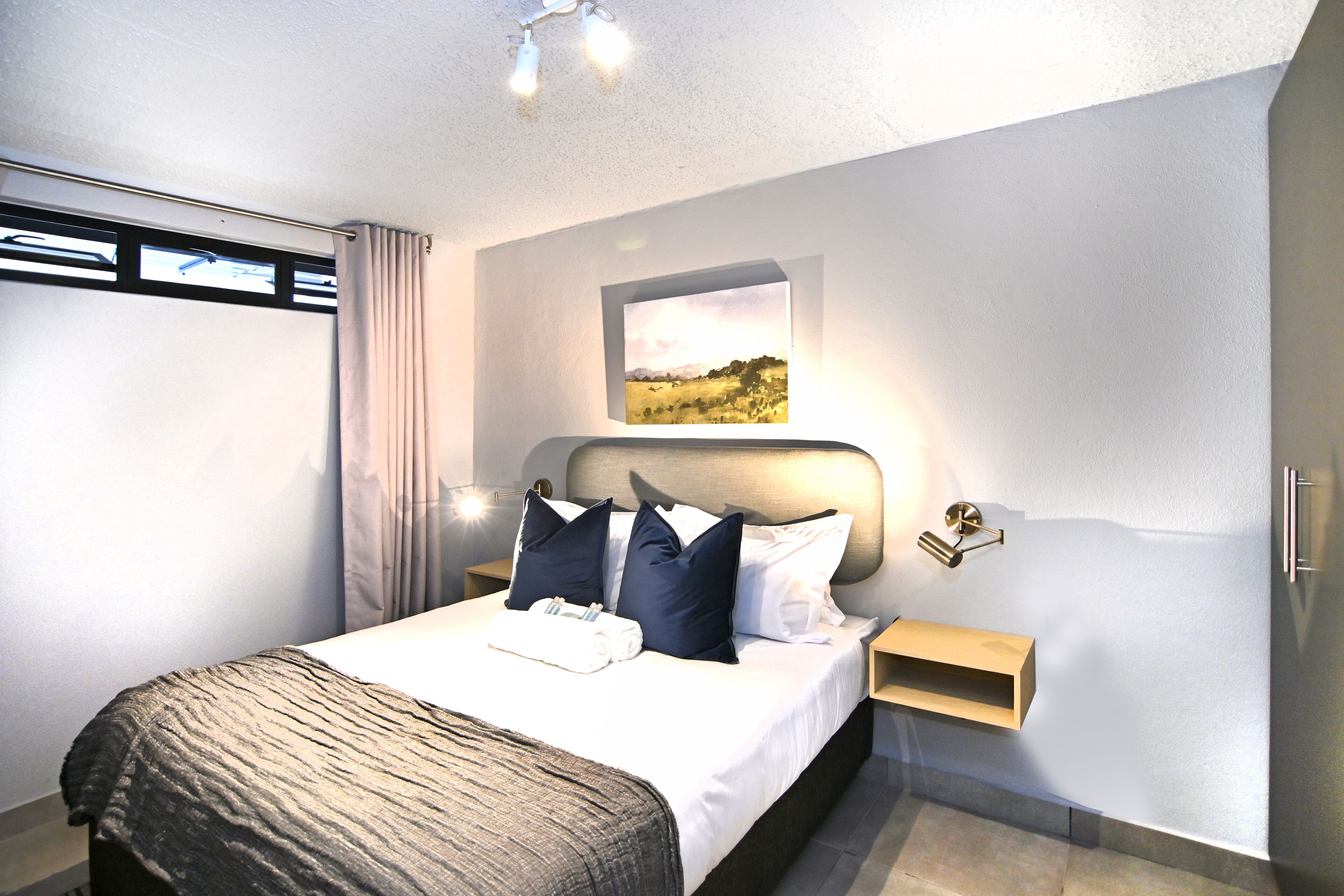 Een moderne hotelkamer met een groot bed, een hoofdbord met geïntegreerde verlichting, een nachtkastje met een lamp, een raam met gordijnen en een ingelijste foto aan de muur.