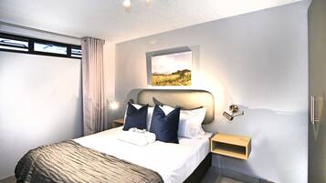 A modern hotel room with a large bed, a headboard with integrated lighting, a bedside table with a lamp, a window with curtains, and a framed picture on the wall.