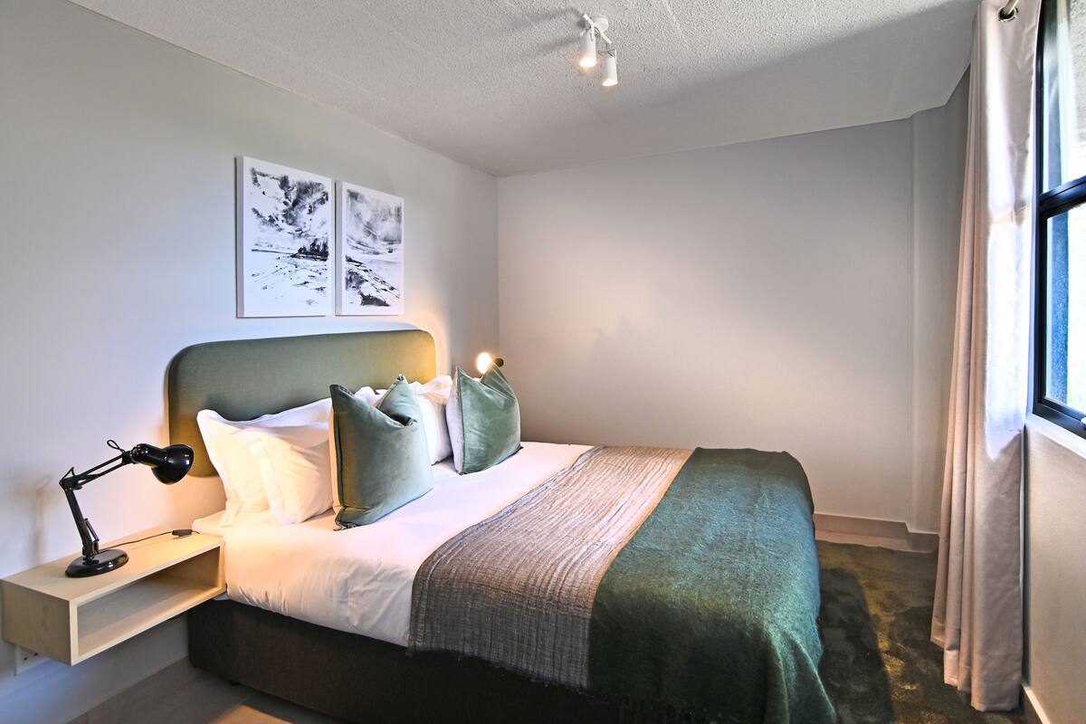 Een net opgemaakt bed met kussens, een bedlampje en een hoofdbord. Boven het bed hangen twee ingelijste foto's aan de muur.