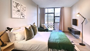 A bedroom with a bed, pillows, a green throw, a nightstand, a lamp, a TV, and a window with curtains.