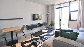 A modern living room with a flat-screen TV, a wooden table, two chairs, a black coffee table, a patterned rug, a sofa with green cushions, and a potted plant.