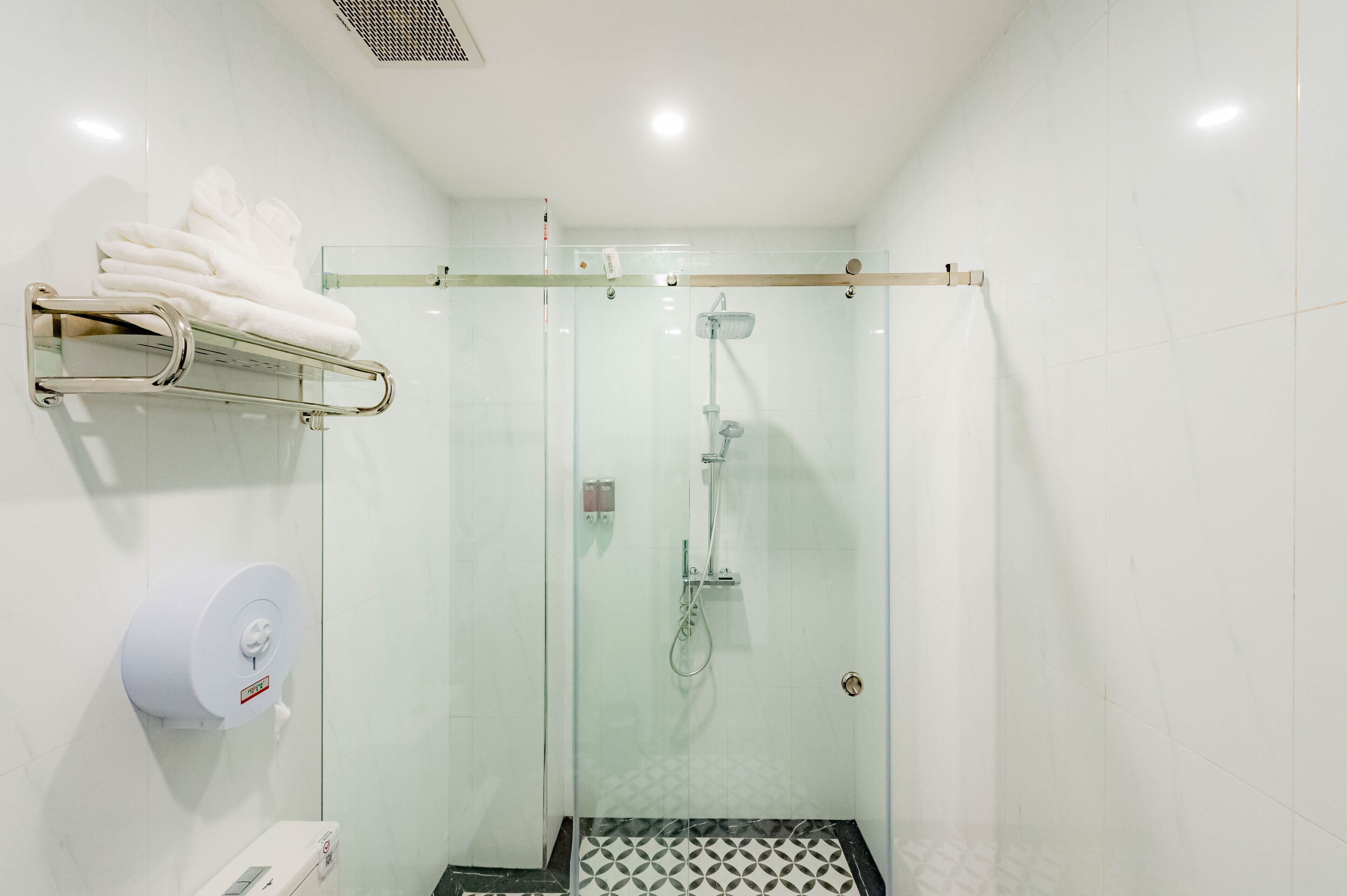 A glass-enclosed shower with a handheld showerhead, a towel rack with folded towels, and a toilet with a paper towel dispenser.