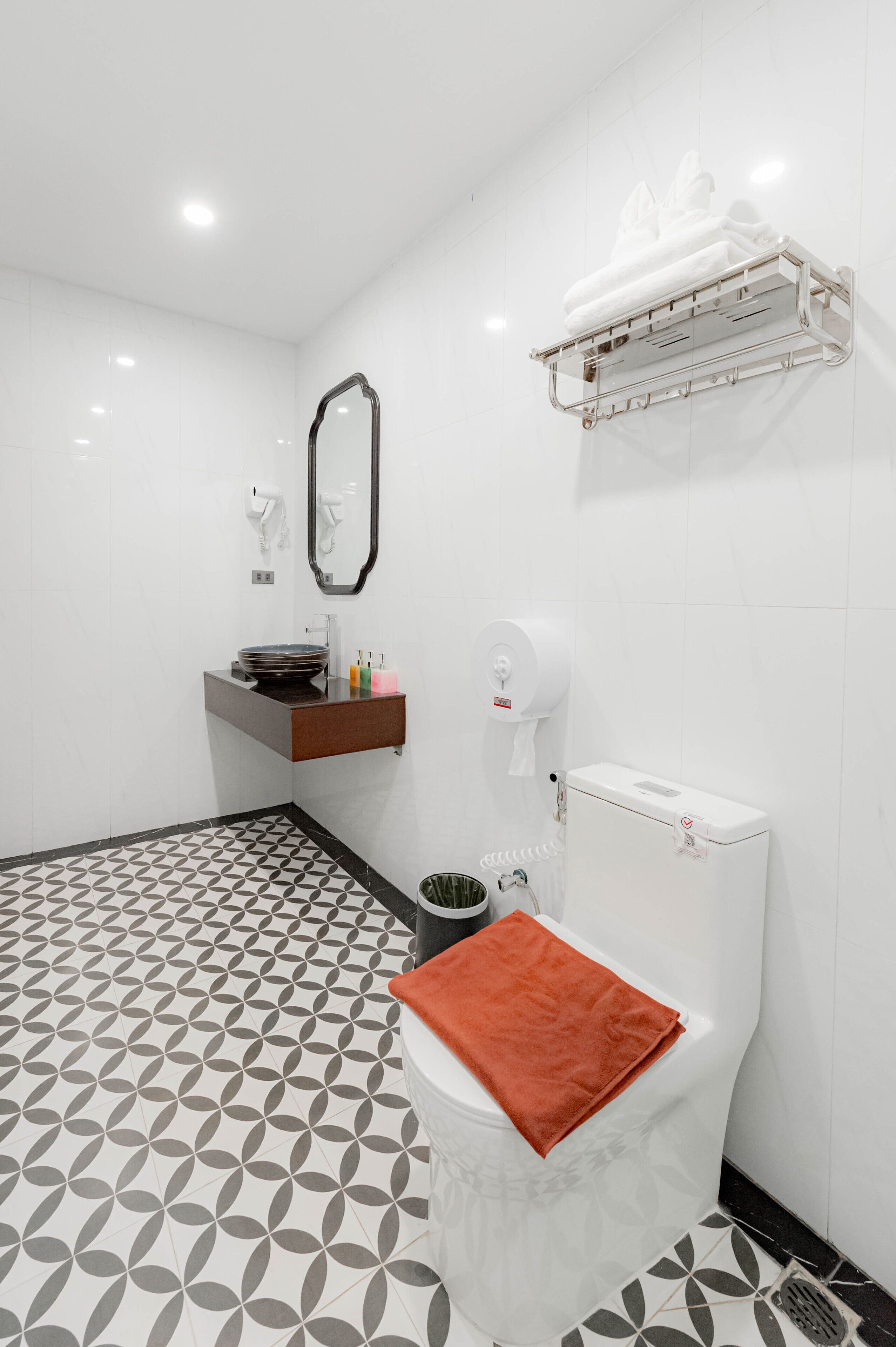 A bathroom with a geometric patterned floor, a white toilet with a red towel, a sink with a mirror above it, and a towel dispenser.