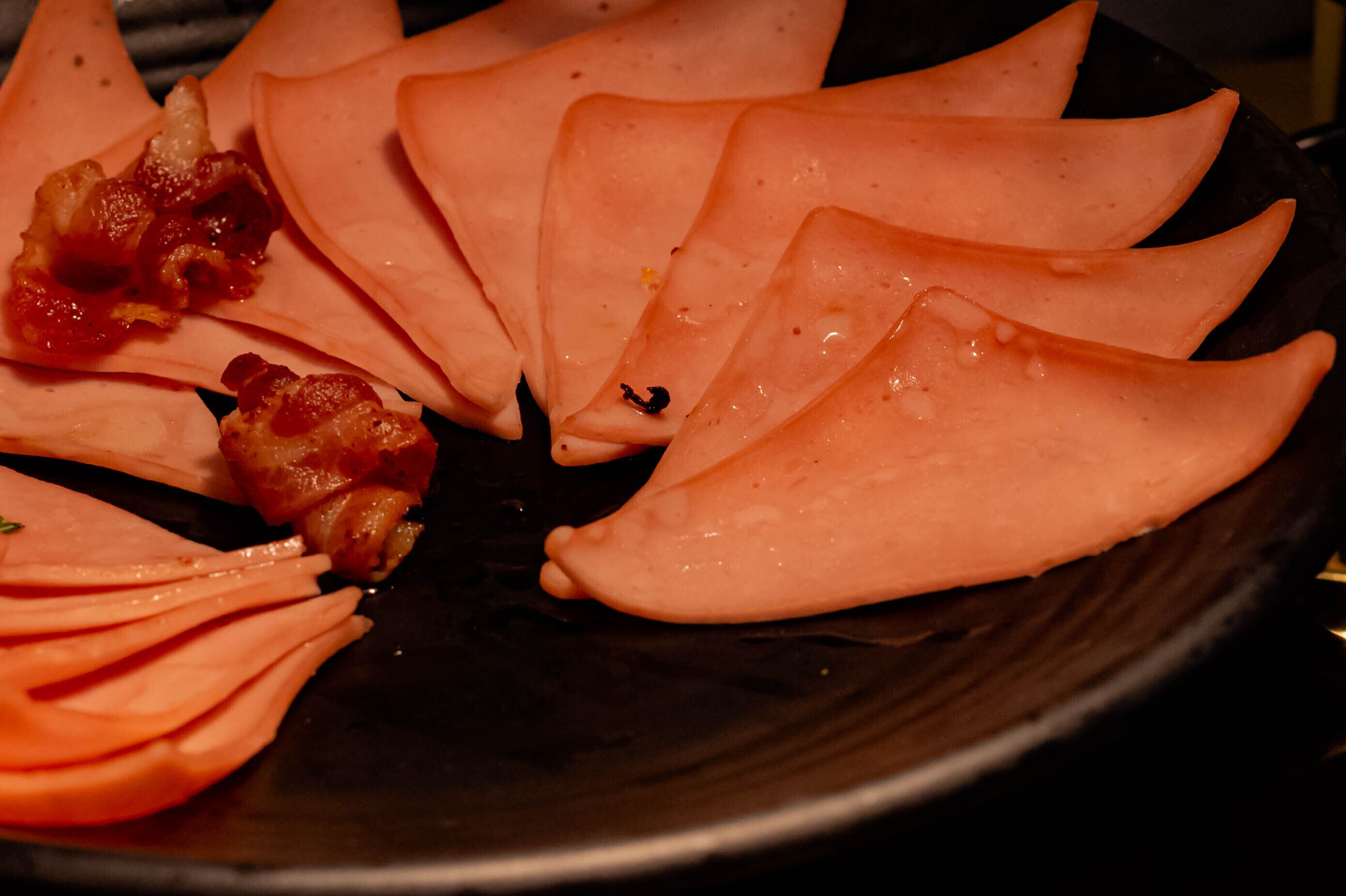 A black plate with neatly arranged slices of pink meat and some bacon pieces.