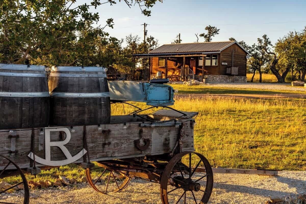 Een houten kar met vaten, een gebouw met een veranda en een grasveld.