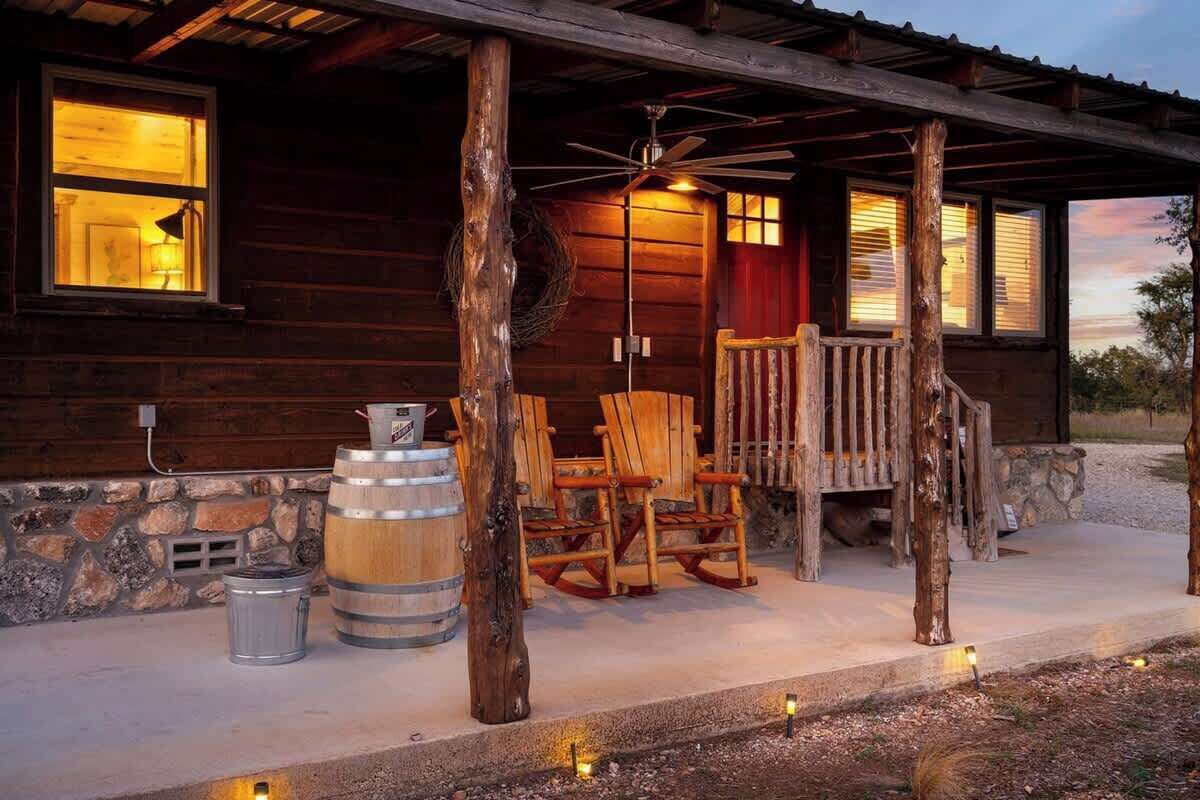 Une cabane rustique dotée d’une véranda, de fauteuils à bascule en bois, d’un tonneau et d’un ventilateur.