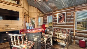 A rustic cabin interior with a wooden dining table, chairs, a television, and a large poster on the wall.