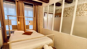A hotel room with a large bed, a wooden bench with towels, a window with curtains, and a stone wall. - Distinguished Townhouse, "L'Aparté" (Verdun)