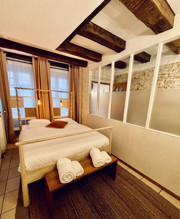 A hotel room with a large bed, a wooden bench with towels, a window with curtains, and a stone wall. - Distinguished Townhouse, "L'Aparté" (Verdun)