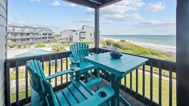 A covered balcony with a view of the beach and ocean, featuring a pool, two chairs, and a table.