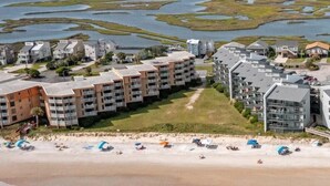 A coastal hotel with multiple-story buildings, a beach with umbrellas, and a marshy area in the background.